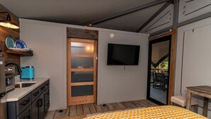 A compact bedroom with a bed, a TV, a small kitchenette, and a sliding door leading to an outdoor area.