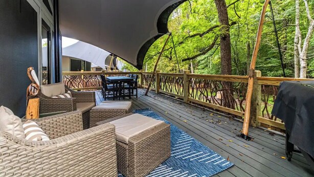 An outdoor deck with wicker furniture, a table, and chairs, surrounded by a wooden railing and trees.