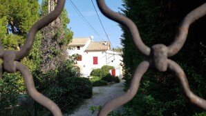 A pathway leading to a white building with red shutters, surrounded by greenery and a metal fence. - Le Guiguet - Mas provençal avec piscine pour 10 (Vedene)