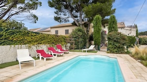 A rectangular swimming pool with a tiled edge, surrounded by a stone deck. There are four lounge chairs with red cushions, two white, and two red, arranged in a row along the pool's edge. - Le Guiguet - Mas provençal avec piscine pour 10 (Vedene)