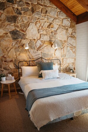 A bedroom with a stone wall, a bed with white and blue bedding, two bedside lamps, and a small table with a plant. - Langmeil Cottages - Wattle (Tanunda)
