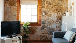 Un salon avec un mur en pierre, une fenêtre, un téléviseur, un canapé, un tapis et un ventilateur de plafond.