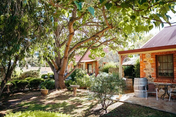 A stone house with a covered outdoor area, surrounded by lush greenery and a large tree. - Langmeil Cottages - Wattle (Tanunda)