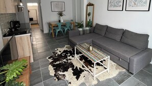 Una sala de estar moderna con un sofá gris, una mesita de centro y un tapete de cuero de vaca. Hay dos cuadros enmarcados en la pared, una zona de comedor con una mesa y sillas, y una cocina con estufa.