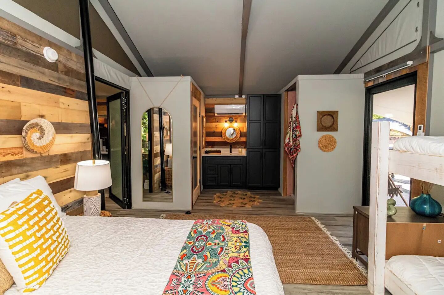 Una habitación de hotel moderna con cama, lámpara de noche, camino de cama colorido, alfombra pequeña, espejo y vista al área de la cocina.