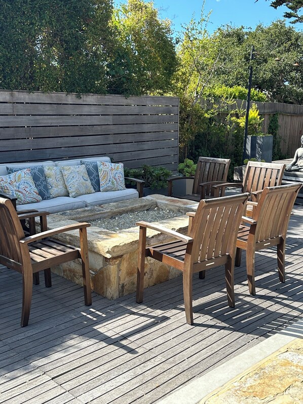 Een houten terras met een vuurkuil van steen, een bank met decoratieve kussens en houten stoelen.