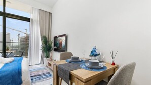 Quarto de hotel moderno com uma janela grande, uma mesa de jantar posta com pratos e copos, uma cama com manta azul e uma planta em vaso.
