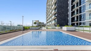 A modern outdoor swimming pool with a wooden deck, surrounded by a high-rise building with balconies. - Charming Chalet Escape (Dubai)