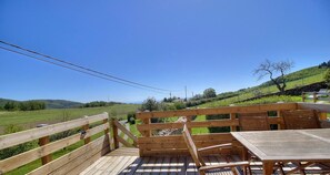 Une terrasse extérieure en bois, avec une table et des chaises, donnant sur un paysage verdoyant avec des collines et un ciel dégagé.