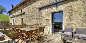 Un bâtiment en pierre doté d’une terrasse en bois, d’un espace extérieur aménagé pour s’asseoir et d’une porte donnant sur une zone abritée.