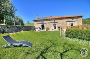 Une maison en pierre avec un toit recouvert de tuiles rouges, une cour verdoyante et deux fauteuils de jardin.