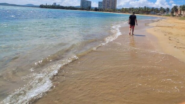 Une plage avec une eau claire, une personne qui se promène et des bâtiments en arrière-plan.