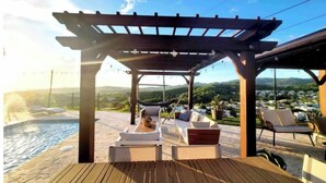 Une terrasse en bois avec des chaises longues et une pergola donnant sur une piscine et un paysage pittoresque.