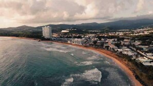 Vista aérea de un pueblo costero con edificios a lo largo de la playa, una extensa masa de agua y colinas a lo lejos.