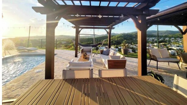 Terraza de madera con sillones de descanso y una pérgola con vistas a una piscina y un paisaje pintoresco.
