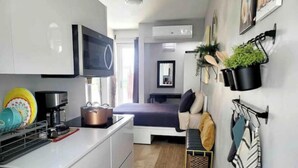 Een compacte keuken met een magnetron, koffiezetapparaat en een klein fornuis. Er is een bed met een blauwe deken en kussens, en een klein bankje met een handdoek. Een potplant hangt aan de muur, en er hangt een spiegel boven het bed.