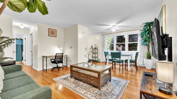 Un salon spacieux avec une table basse en verre, un coin repas avec une table blanche et des chaises turquoise, et un téléviseur fixé au mur.