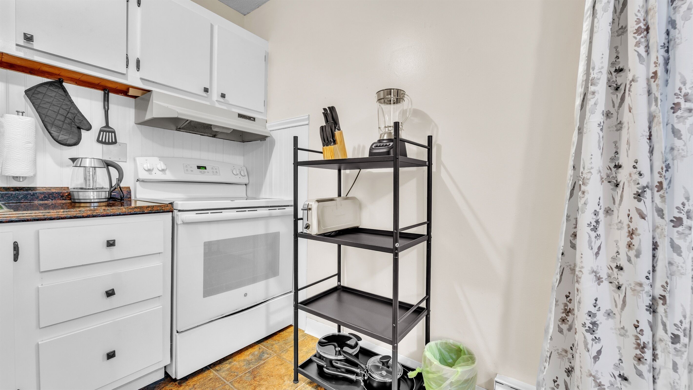 A compact kitchen with white cabinets, a stove, a kettle, and a small shelf with kitchen utensils.