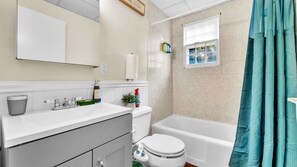 Une salle de bain avec un lavabo, des toilettes et une baignoire, tous de couleur blanche. Une fenêtre offre une vue sur les arbres à l’extérieur.