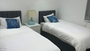 Ein Hotelzimmer mit zwei Betten, weißer Bettwäsche, einem Nachttisch mit Lampe und einem Fenster mit Jalousien.
