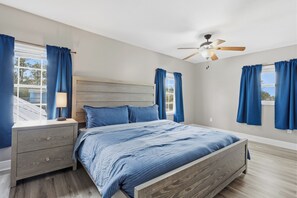 Une chambre à coucher avec un cadre de lit en bois, une table de chevet, un ventilateur de plafond et de grandes fenêtres ornées de rideaux bleus.