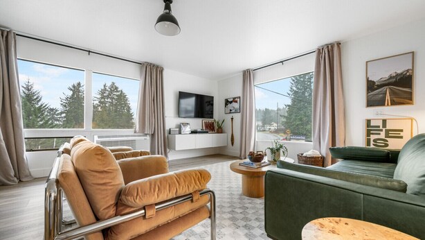 A modern living room with a sofa, armchair, and a coffee table. There is a TV mounted on the wall, and a window with curtains. - Luxury Harbor Escape | Walk to Food & Fun (Juneau)