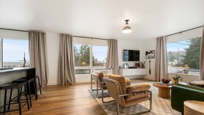 A spacious living room with a wooden floor, a sofa, a chair, a TV, and a window with curtains. - Luxury Harbor Escape | Walk to Food & Fun (Juneau)