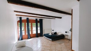 A bedroom with a bed, a desk, and a chair. There is a window with curtains and a view of greenery outside.