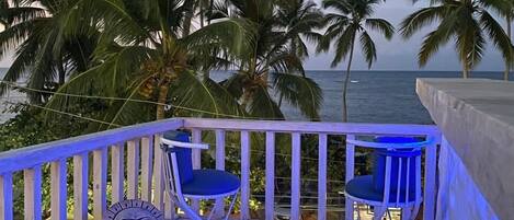Un balcón con dos sillas y una escultura decorativa, con vista a una playa con palmeras y el océano.