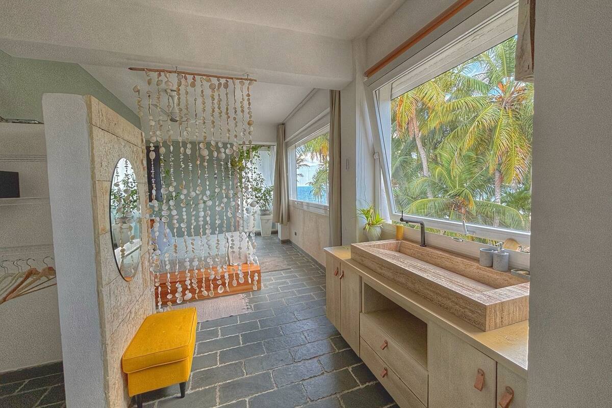 A modern bathroom with a stone countertop, a yellow bench, and a view of palm trees through a large window.