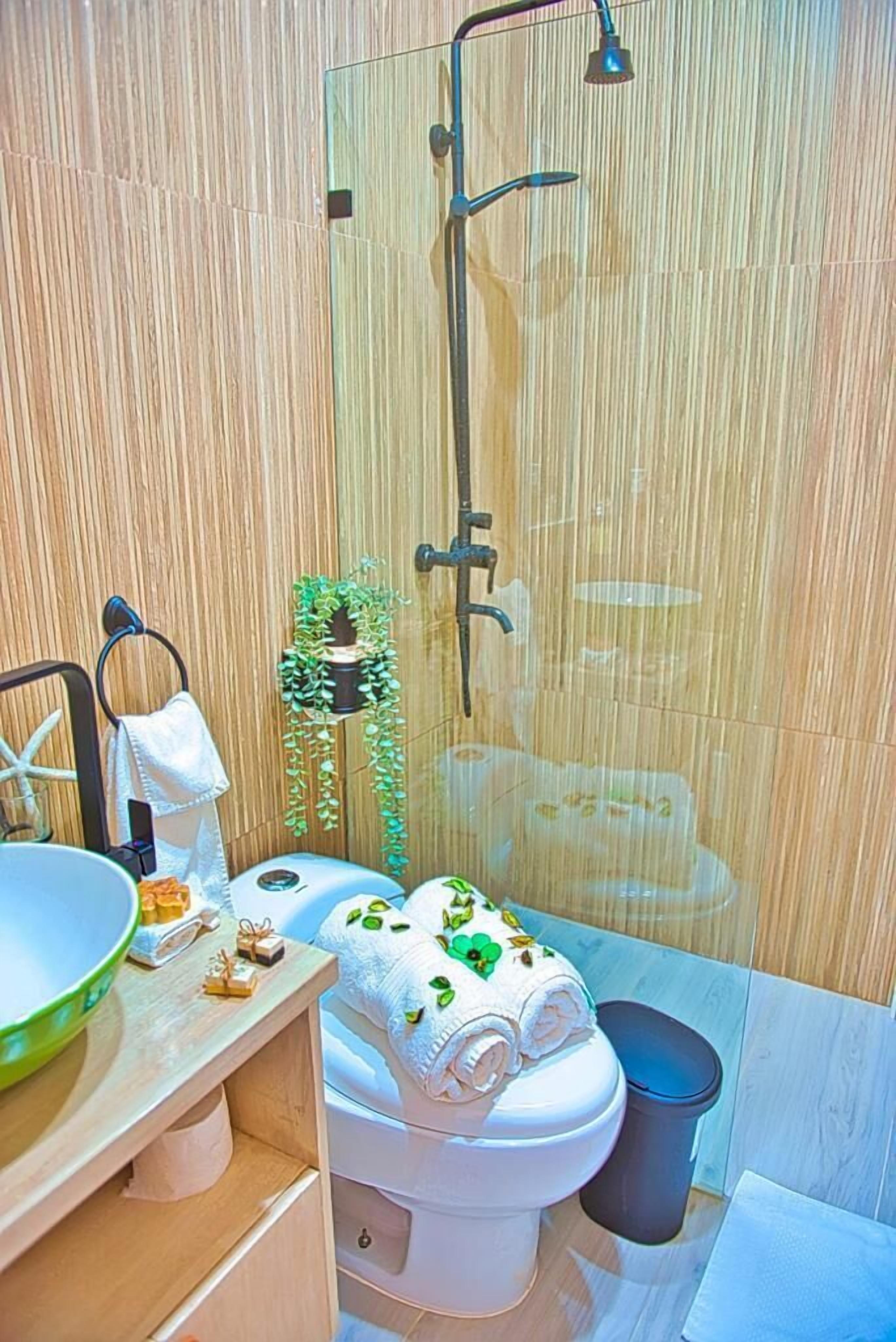 A bathroom with a wooden wall, a glass shower enclosure, a toilet, a sink with a bowl, and a trash bin.