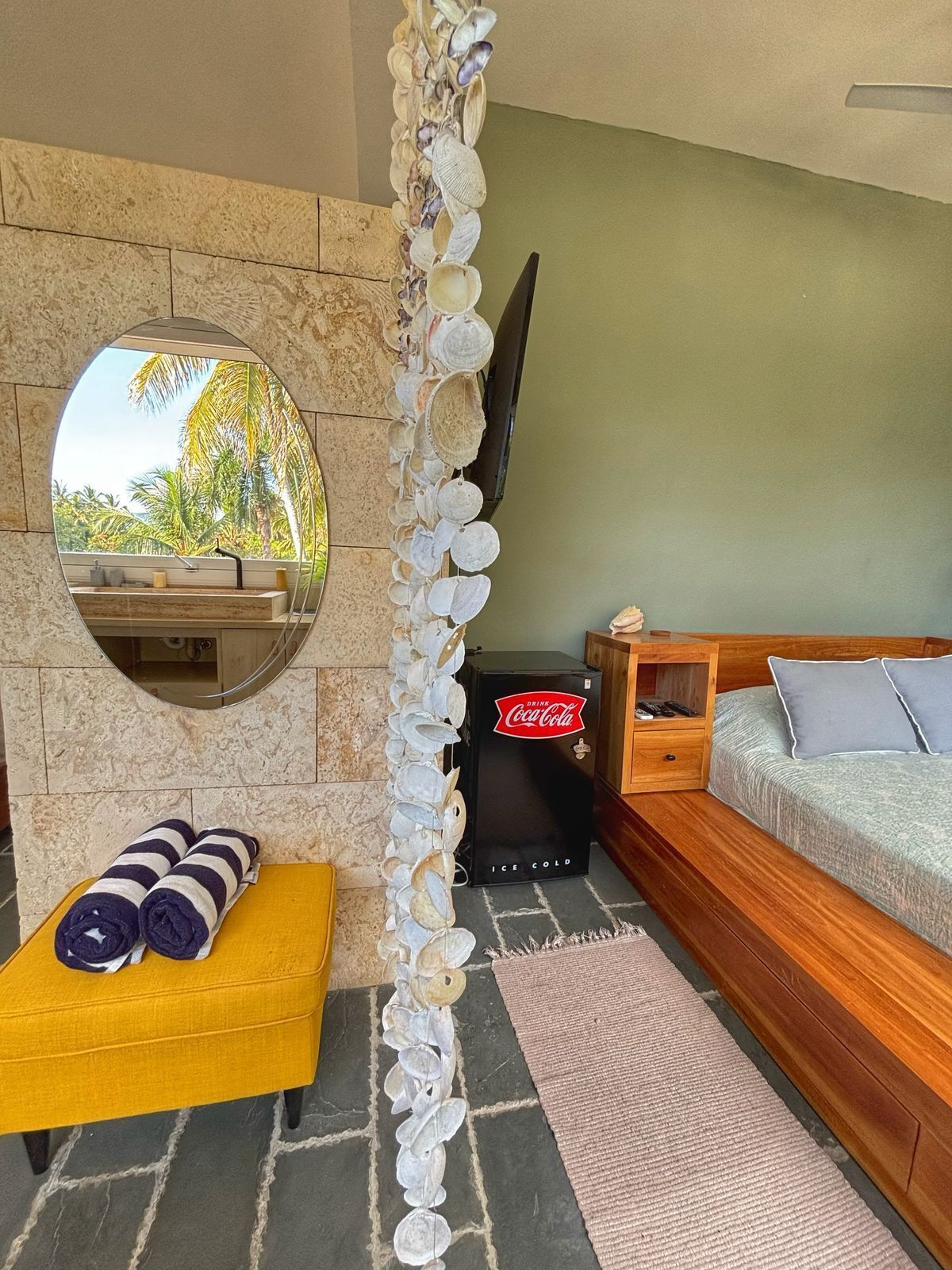 A cozy room with a stone wall, a round mirror, a wooden sofa, a yellow ottoman, and a Coca-Cola refrigerator.