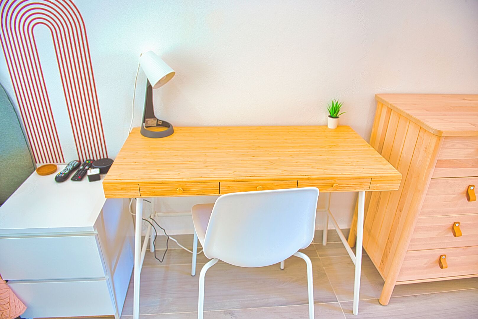 Un bureau en bois avec une lampe, une chaise blanche, une petite plante et un meuble en bois avec des tiroirs.