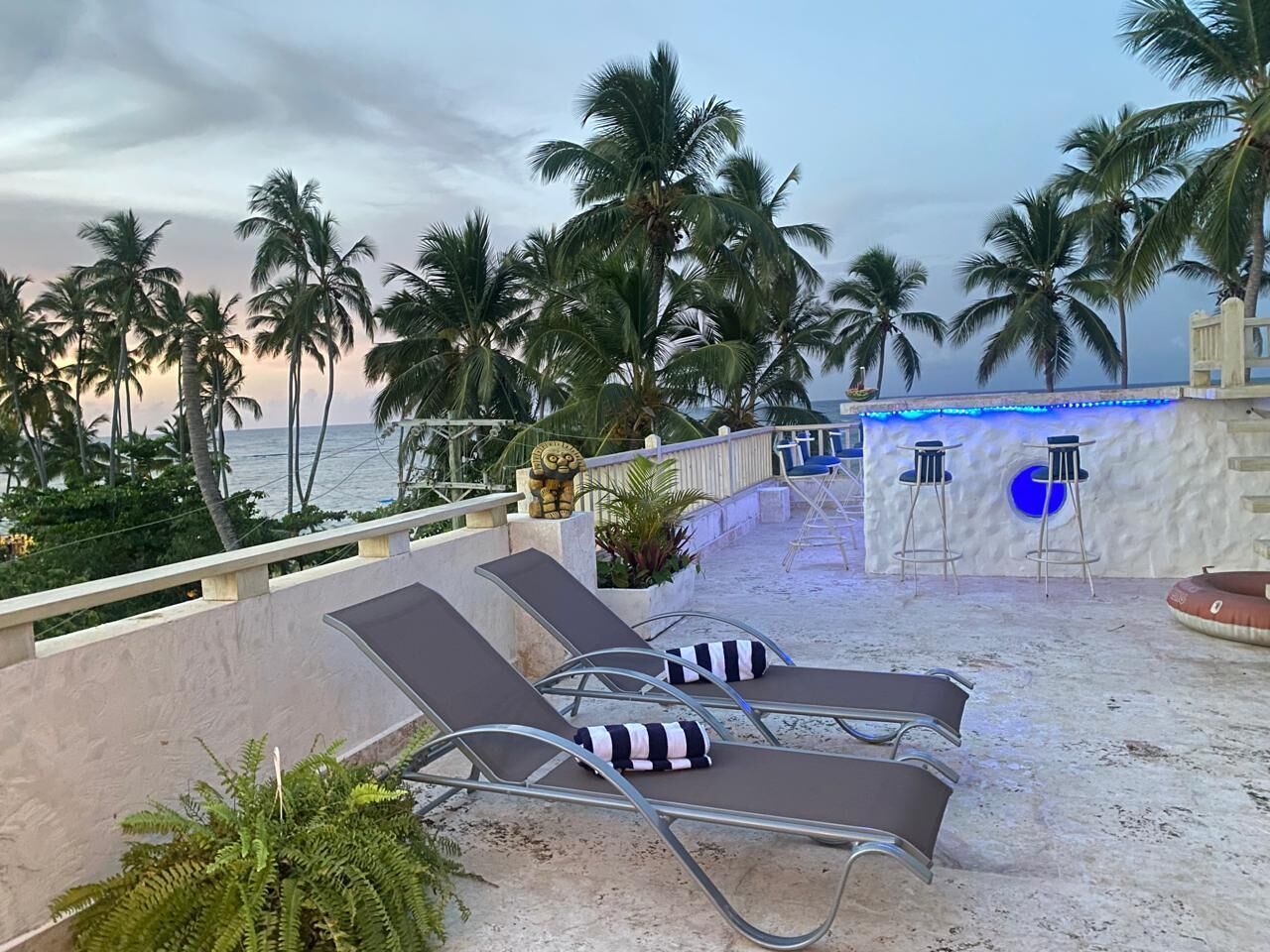 A beachside lounge area with sun loungers, a bar with blue lighting, and palm trees in the background.