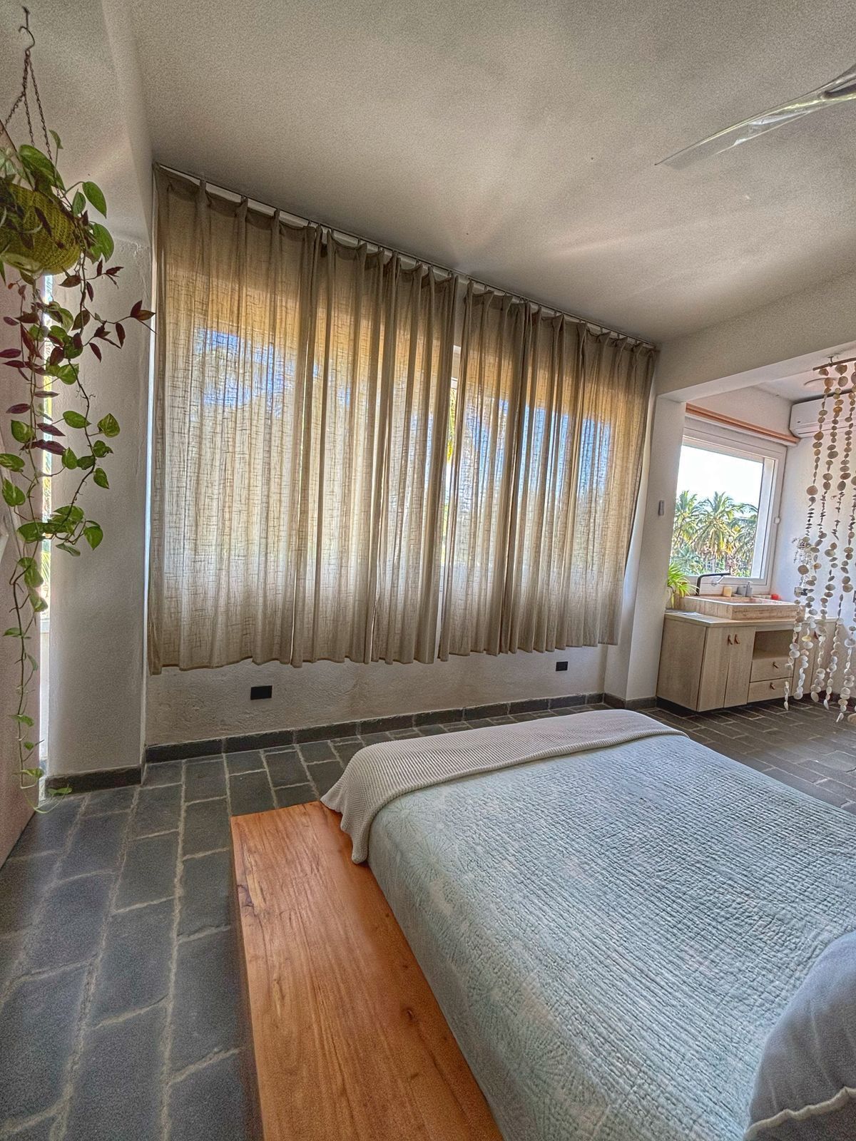A bedroom with a large bed, a wooden bench, a window with curtains, a small kitchenette, and a hanging plant.