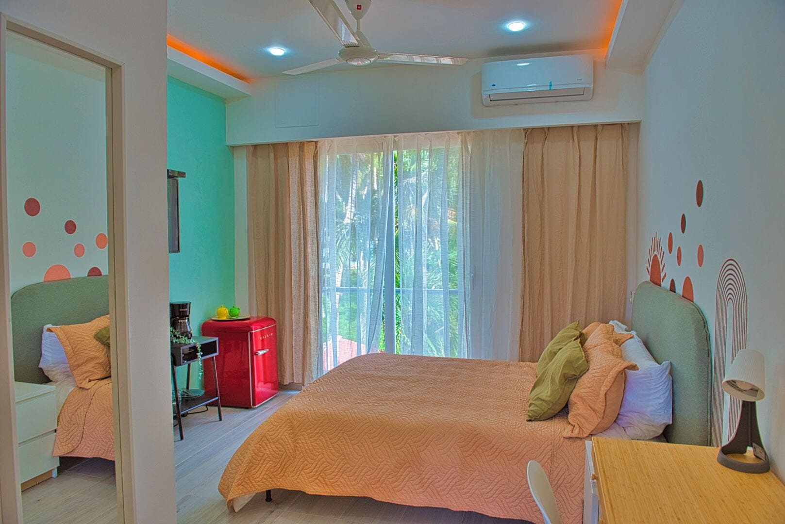 A bedroom with a large bed, a desk with a chair, a red refrigerator, and a ceiling fan.