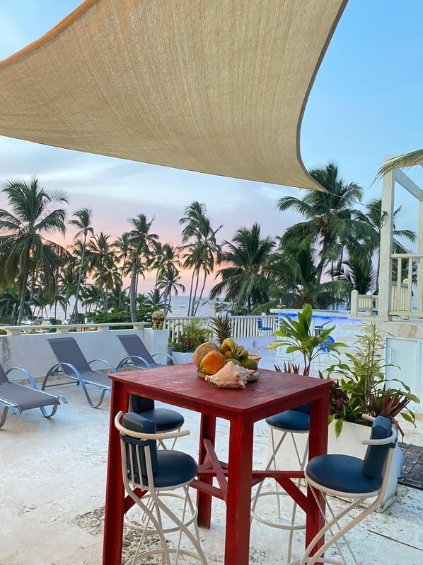 A red table with four blue chairs and a tablecloth, surrounded by a shaded area with a view of palm trees and a pool. - MARINA BAY HOTEL BOUTIQUE (Las Terrenas)