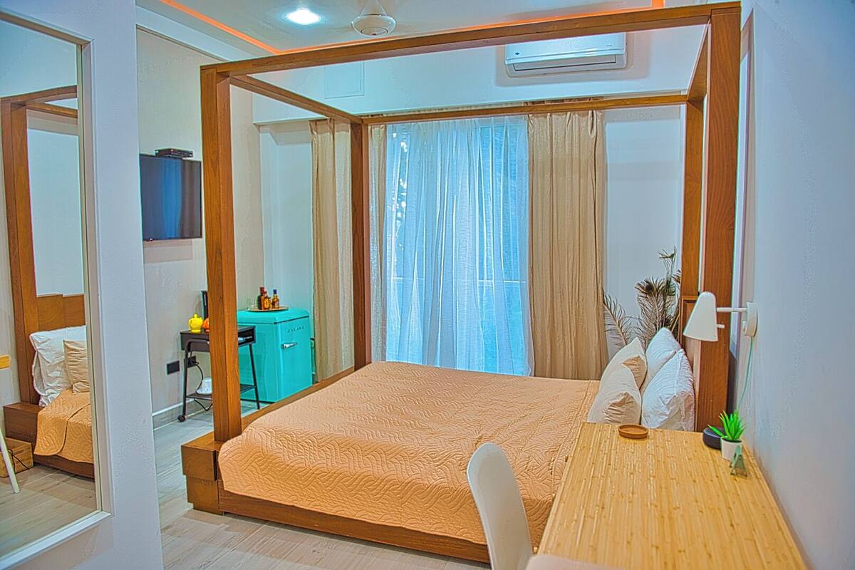 A bedroom with a bed, a desk, a chair, a small table, and a potted plant.