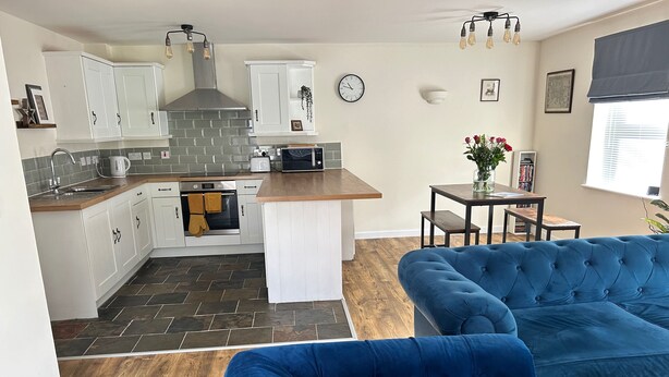 A modern kitchen with white cabinets, a built-in oven, and a microwave. There is a blue velvet sofa in the living area.