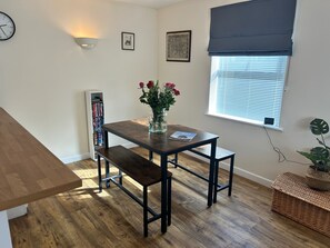 Un coin repas avec une table en bois, des bancs, un vase de fleurs, une bibliothèque et une fenêtre avec des stores.