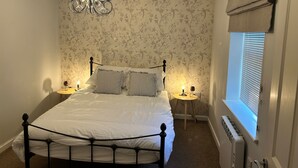 A bedroom with a four-poster bed, two bedside tables with lamps, a chandelier, floral wallpaper, and a window with blinds.