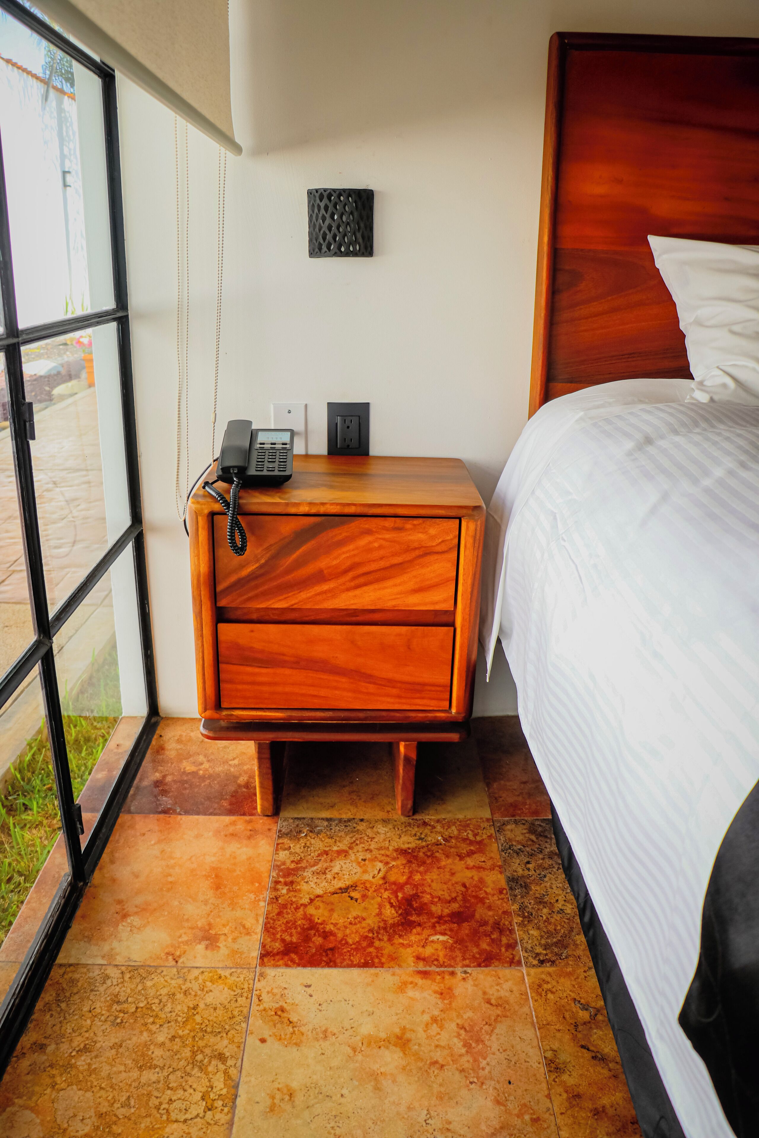 Una habitación de hotel con una mesita de noche de madera, un teléfono, un enchufe y una cama con ropa blanca.
