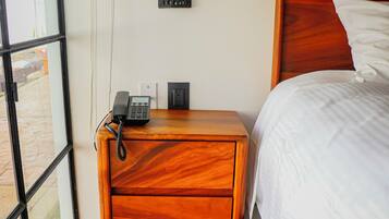 Una habitación de hotel con una mesita de noche de madera, un teléfono, un enchufe y una cama con ropa blanca.