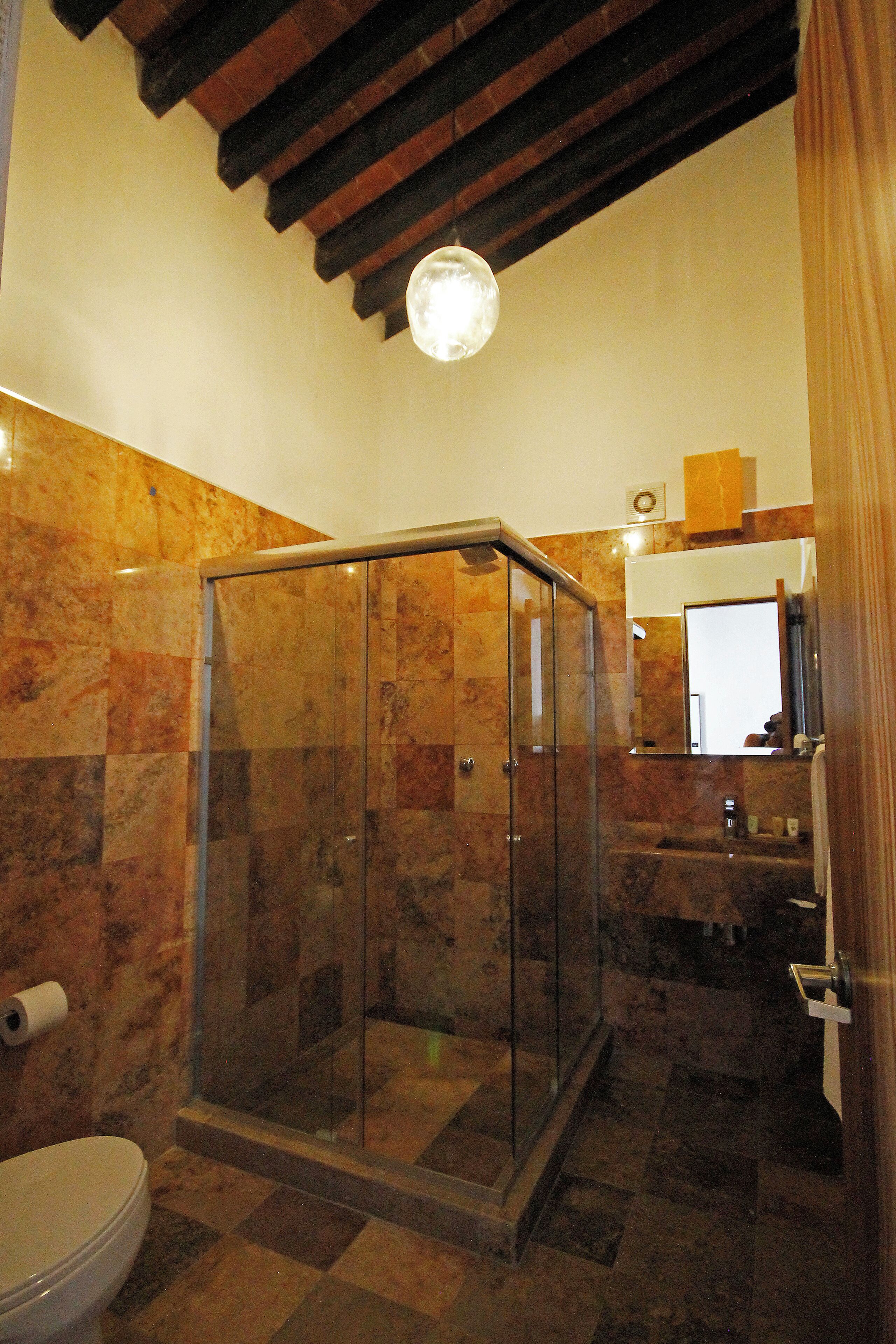 Un baño con ducha de mampara de vidrio, paredes de mármol, inodoro y espejo sobre el lavamanos.