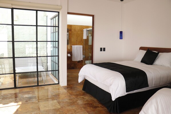 Quarto de hotel com uma grande porta de vidro deslizante que dá para uma varanda, uma cama com roupa de cama branca e detalhes em preto, e uma pequena luminária pendente azul.