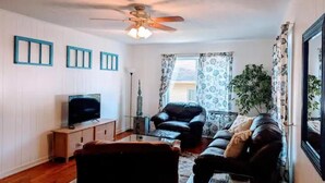 A living room with a ceiling fan, a television on a wooden stand, two leather sofas, a glass coffee table, and a large potted plant.