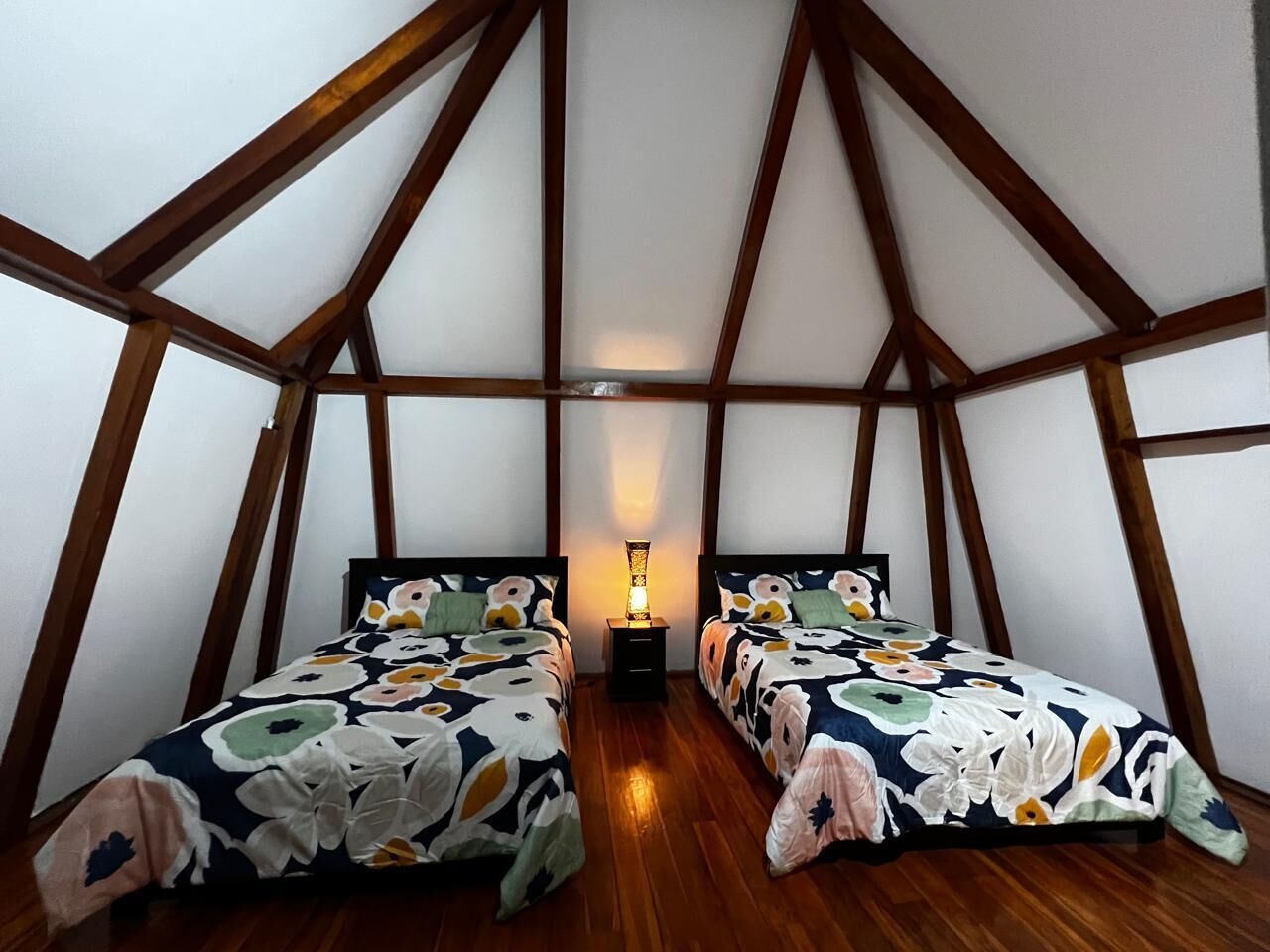 A small, triangular attic bedroom with two beds, wooden beams, and a nightstand with a lamp.