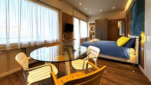 Une chambre d’hôtel moderne avec une table ronde en verre, des chaises blanches, un lit avec des coussins bleus et jaunes, une télévision et une grande fenêtre avec des rideaux.
