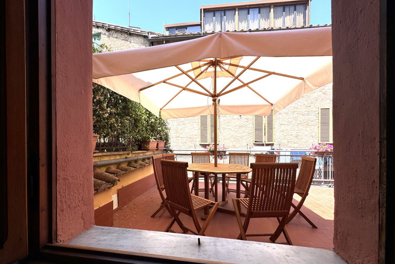 Une terrasse avec une table et des chaises sous un grand parasol, entourée de plantes en pot et d’un mur de pierres.