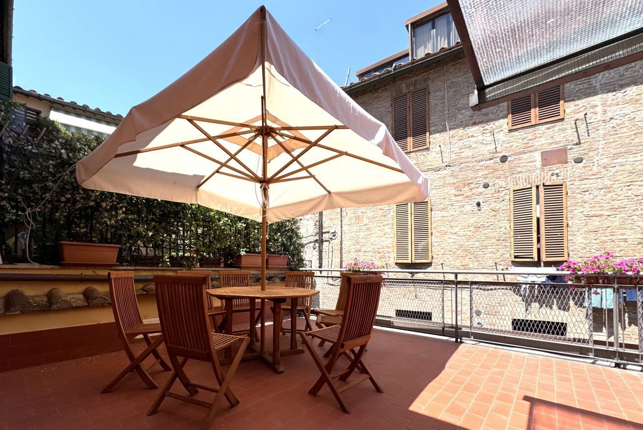 Eine Terrasse im Freien mit Tisch und Stühlen unter einem großen Sonnenschirm, umgeben von einem Gebäude mit Fensterläden.